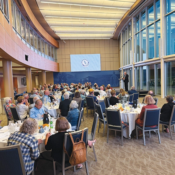 A large gathering of people seated at round tables in Woster Celebration Hall, attentive to a speaker.