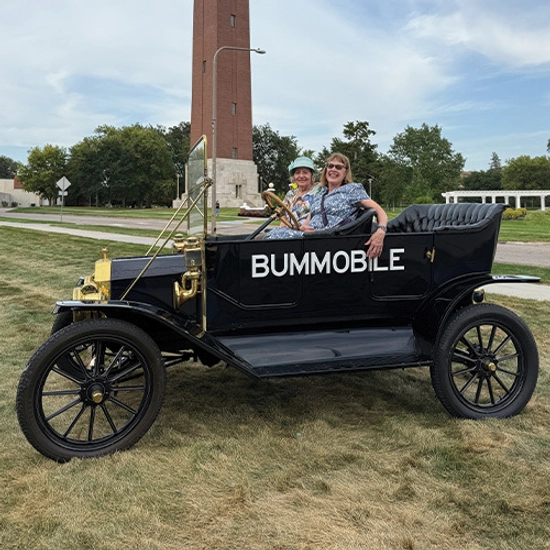 A vintage black car labeled "BUMMOBILE" with two smiling people sits on the grass.