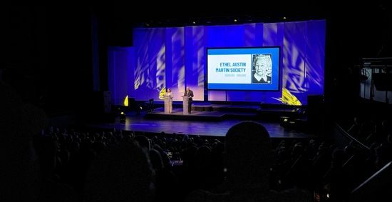 Two presenters stand on stage with a large screen during the Celebration of Philanthropy program.