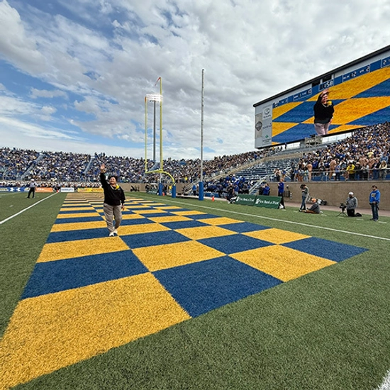 A distinguished alumnus stands in the blue and yellow checkerboard endzone at Dana J. Dkyhouse Stadium, waving to the crowd.