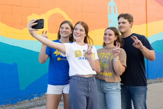 Four people stand smiling and taking a selfie in front of a colorful mural with abstract shapes and a building silhouette.