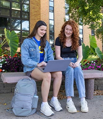 Two young women sit on a bench outside, smiling and looking at a laptop.