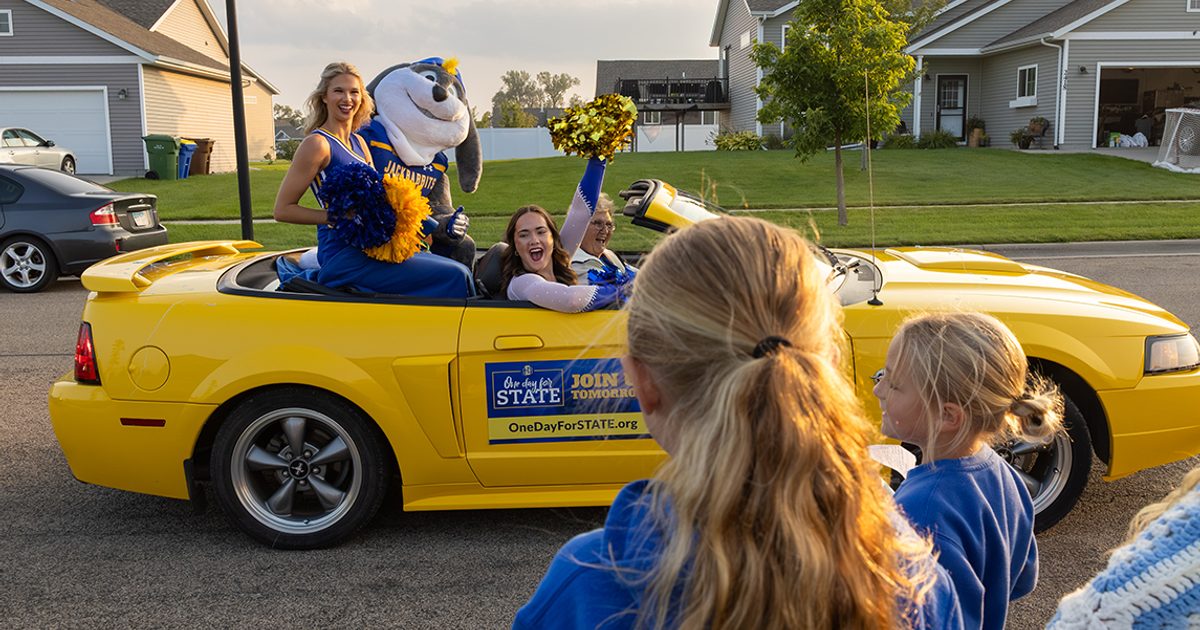 Jack rides in a yellow convertible with cheerleaders, waving at children alongside the road.