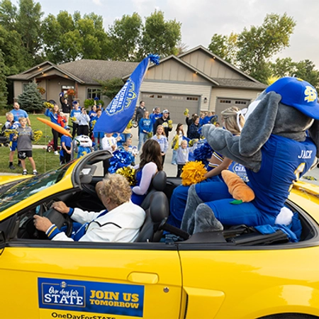 Jack waves at a large group of people standing in a driveway.