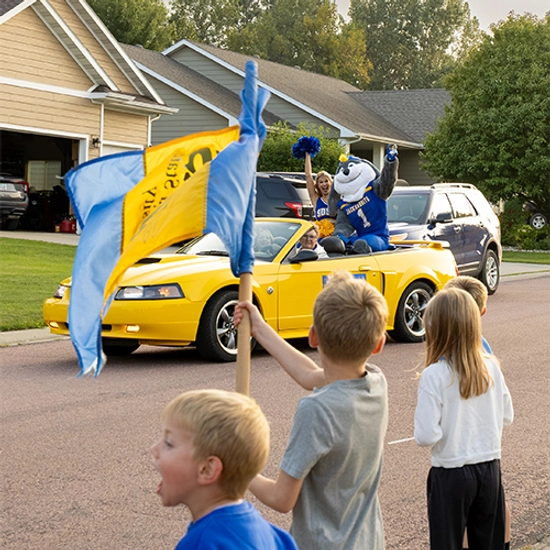 Jack rides in a yellow convertible with cheerleaders, waving at children alongside the road.
