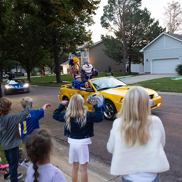 Jack rides in a yellow convertible with cheerleaders, waving at children alongside the road.