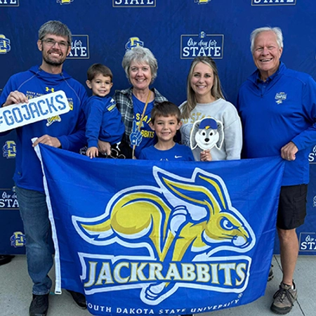 Family of six standing in front of a One Day for STATE banner, all wearing SDSU gear and holding up a Jackrabbit flag.