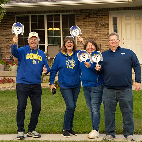 Group of 4 adults wearing Jacks gear, smiling on a sidewalk, and holding up paper Jackrabbit faces.