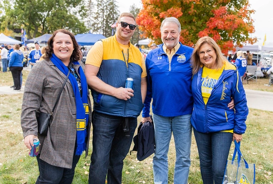 Four people wearing SDSU blue and yellow are smiling for the camera outside the SDSU alumni tailgate tent.