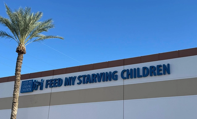 The image shows the exterior of a building with a palm tree and a clear blue sky. A sign reads "FEED MY STARVING CHILDREN" in large blue letters.