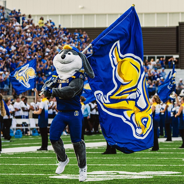 The Jackrabbit mascot stands on the football field, holding a Jackrabbit flag before kickoff.