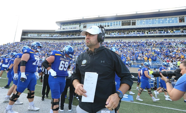 SDSU Football Head Coach Dan Jackson, wearing a headset, stands on the sideline with SDSU football players behind him in the Dana J. Dykhouse Stadium.