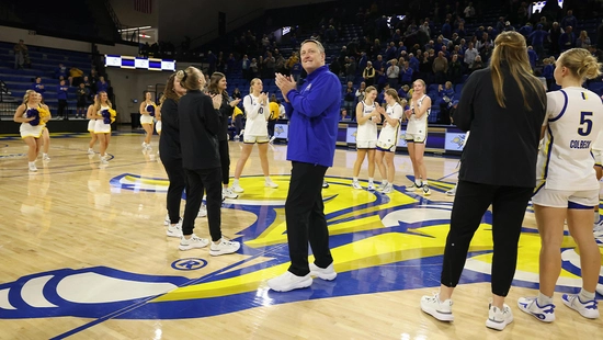 Aaron Johnston standing in the middle of the basketball court at First Bank & Trust arena with women's basketball players standing around him.
