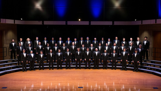 A large choir of men in black tuxedos stands on tiered risers on a wooden stage, under soft blue and white lighting.