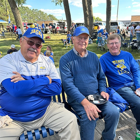 Three smiling men in baseball caps and SDSU shirts sit together outside of the First Bank & Trust Alumni Rabbit Den tailgate tent.