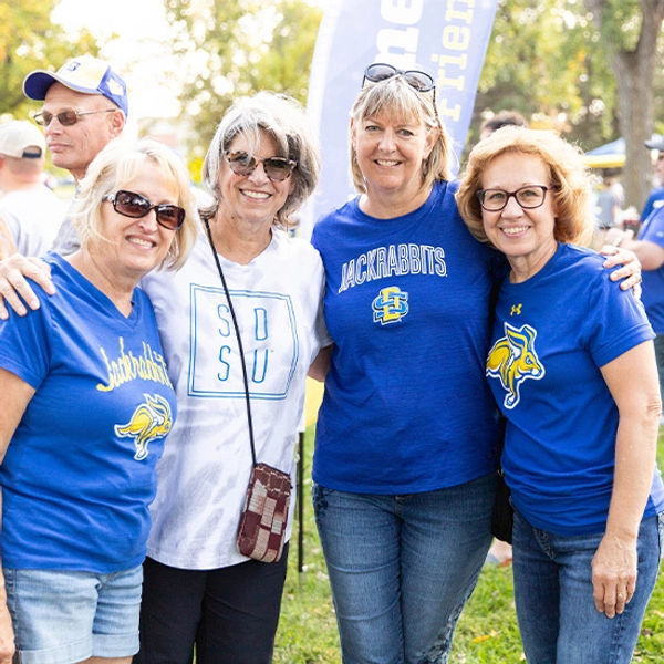 Four smiling women pose outdoors with arms around each other, wearing blue and yellow SDSU shirts outside of the First Bank & Trust Rabbit Den alumni tailgate tent.