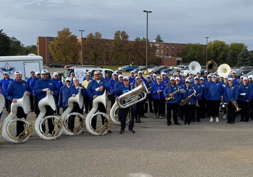 A large group of Pride of the Dakotas Alumni wearing blue coats and white hats posing in a parking lot with instruments before marching in the Hobo Day Parade.