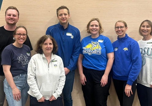 A group of seven SDSU alumni and friends standing in a row, smiling for the camera against a beige wall after a TC Jackrabbits volunteer opportunity.