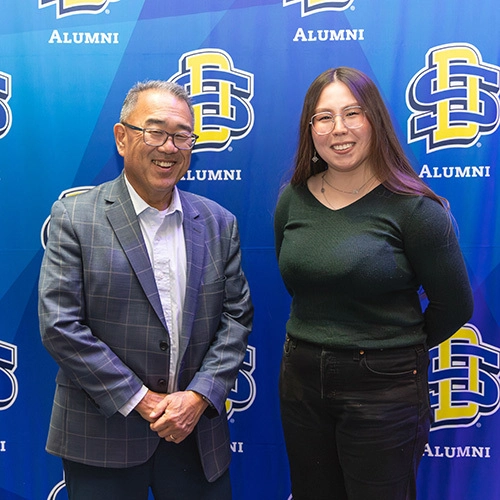 A mentor and mentee pair smiling for the camera in front of a blue SDSU photo backdrop.