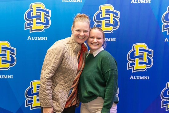 A mentor-mentee pairing smile together with an SDSU Alumni background behind them.