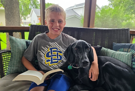 A young boy smiles and sits on a porch holding a book open, with his arm around a black dog.