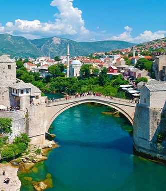 A historic stone bridge arches over a turquoise river, surrounded by lush greenery and charming red-roofed buildings under a bright blue sky.