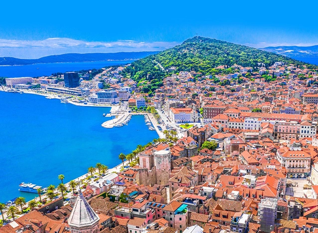 Aerial view of a coastal city with terracotta rooftops, a turquoise harbor, and lush green hills under a clear blue sky.