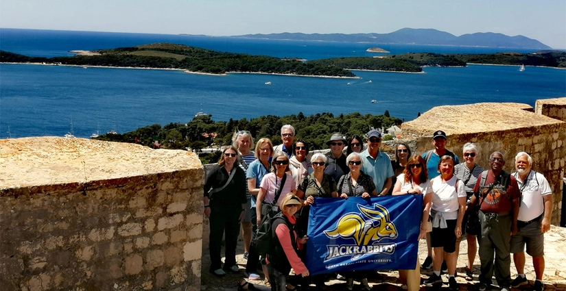 A large group of people traveling together pose for a group photo in front a beautiful, large body of water during their Jackrabbit Journeys trip.