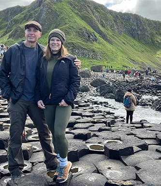 Two people smile side by side at the Giant's Causeway with beautiful scenery around and behind them.