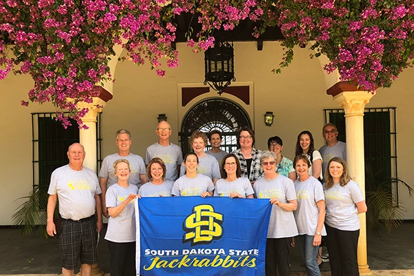 A large group of Jackrabbits traveling together pose for a group photo in Spain with a flower arch above them.