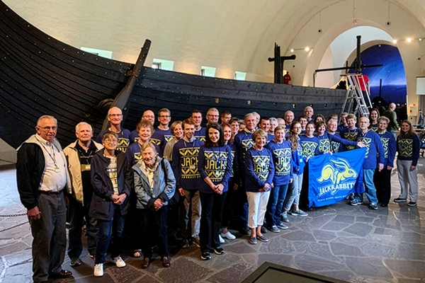 A large group of Jackrabbits traveling together pose for a group photo in Norway with a Vikings ship behind them.
