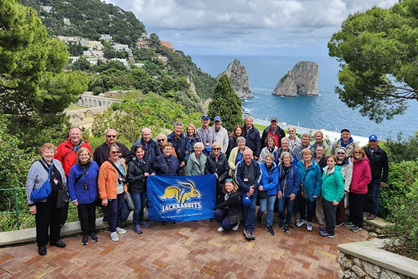 A large group of Jackrabbits traveling together pose for a group photo in Greece with a coastline in the background.