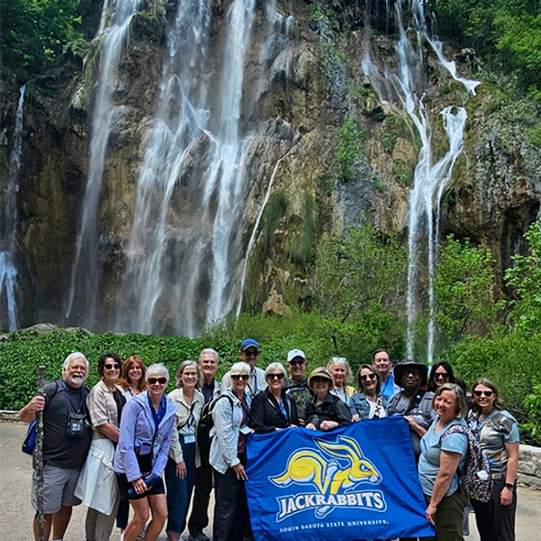 A large group traveling with Jackrabbit Journeys smiles together in front of a beautiful waterfall.