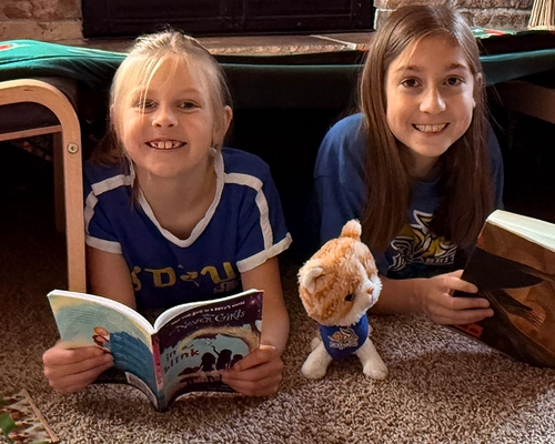 Two young girls in a blanket fort reading books.