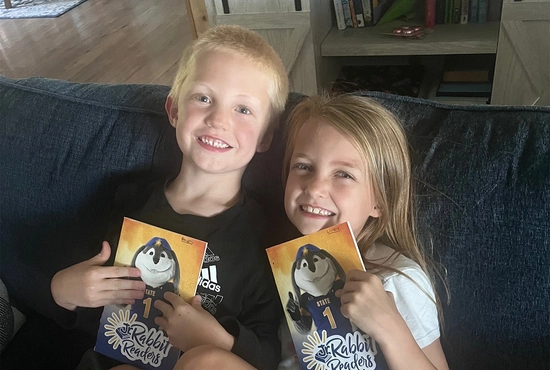 Two children smile together on a couch, holding Jr. Rabbit Reader booklets.