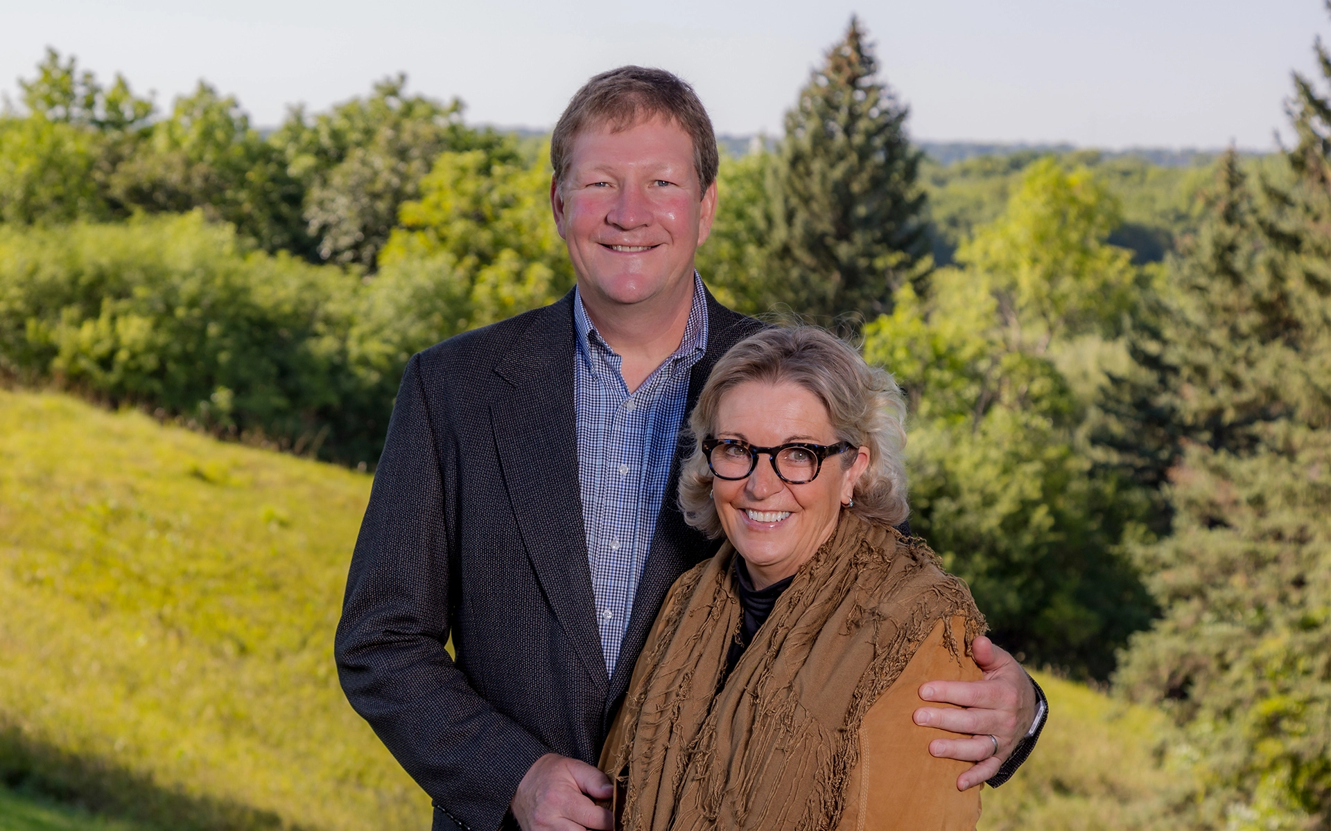 Outdoor photo of Brian and Denise Aamlid, smiling next to one another while Brian has his arm around Denise.