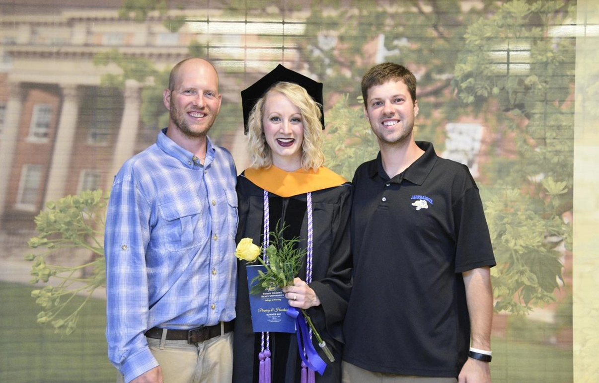 Molly's husband and Dane Yde stand alongside Molly, dressed in her commencement ceremony cap and gown for her Doctor of Nursing Practice (DNP) degree.