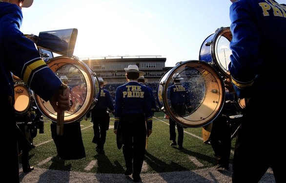 Pride of the Dakotas walking on the football field at the Dana J. Dykhouse stadium, with the sun just peeking over the stands and the drum rims lit up from the glow.