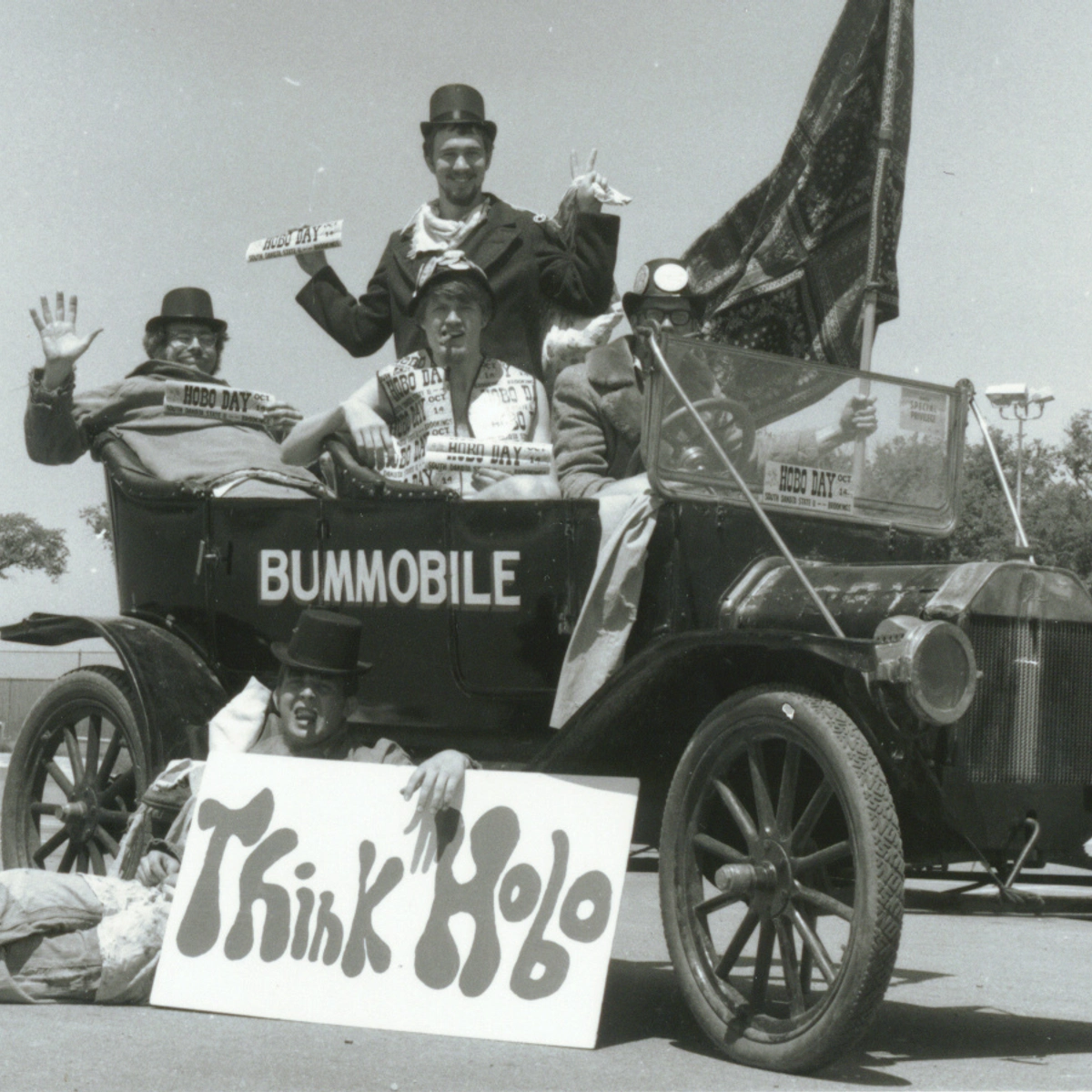 Historic photo of members of the hobo day committee in the Bummobile with members holding a sign that says "think hobo"