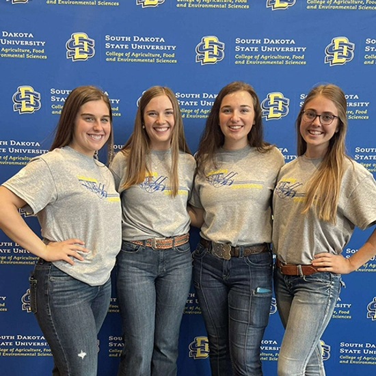 Abby smiles with three other female students infront of a SDSU themed step and repeat banner.