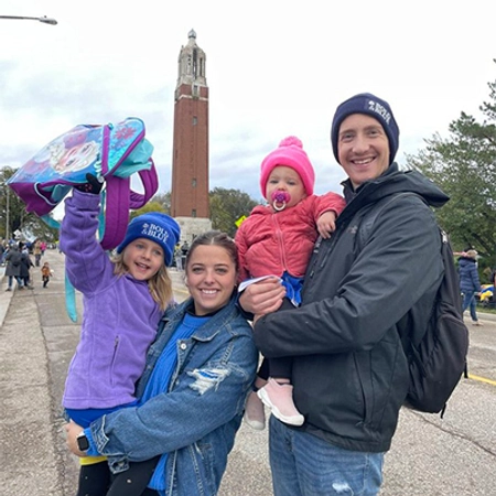 Allison and her husband Woody holding their two daughters, all smiling, with the Hobo Day parade and the SDSU Campanile in the background.