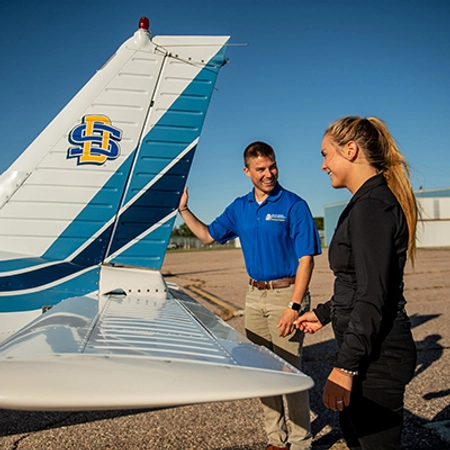 Two SDSU aviation students stand near the tail of an airplane, adorned with blue strips and the SDSU logo.