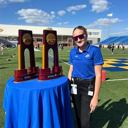cadence koenig smiles next to the two ncaa fcs football championship trophies while on the football field