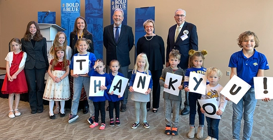 Tate Profilet and Mary DeJong pose with Dr. Carie Green, her children, and President Dunn during the investiture ceremony. Eight children stand in front of them, holding up letters spelling out "thank you!"