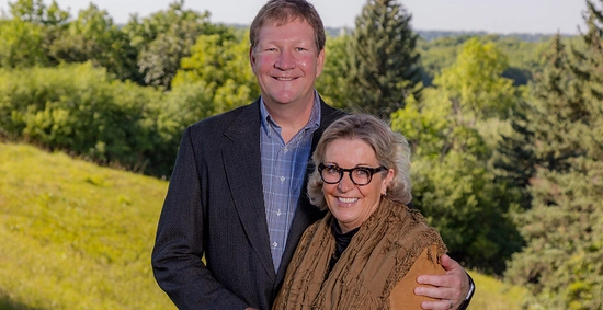 Outdoor photo of Brian and Denise Aamlid, smiling next to one another while Brian has his arm around Denise.