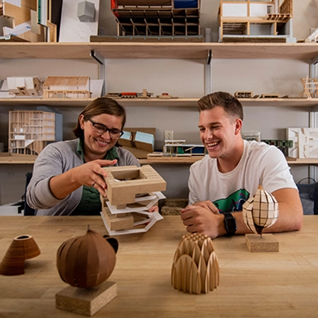 Female architecture professor and a male student smile while looing over a building model.