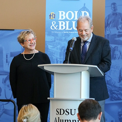 Tate Profilet speaks to the investiture crowd as Mary looks at him, smiling.