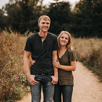 Sydney and Nathan Adrian smile at the camera while taking engagement photos in a field of browning grass.