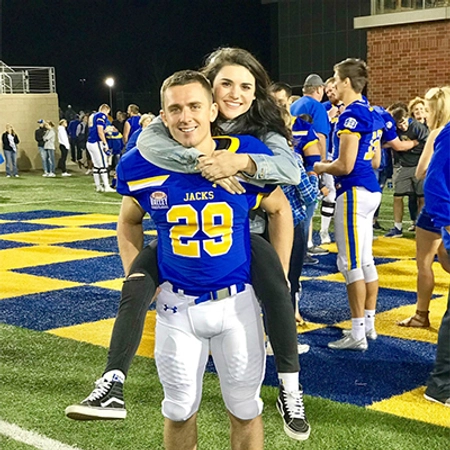 Hanna Eide getting a piggyback ride and smiling with her husband on the SDSU football field.