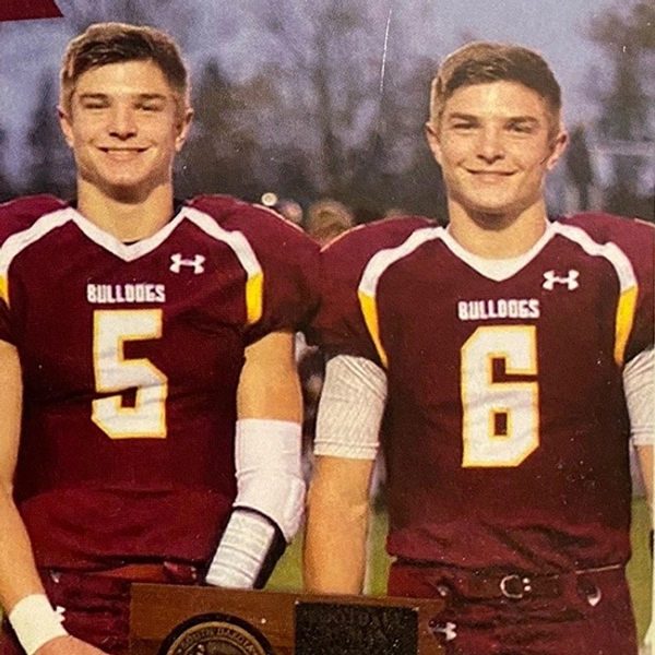 Janke twins standing side by side during their high school years in their football uniforms on the field, holding a football trophy